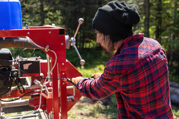 Lumberjack is seen from behind wearing black hat and red checked shirt, operating an industrial saw in woodland with blurry trees in background.