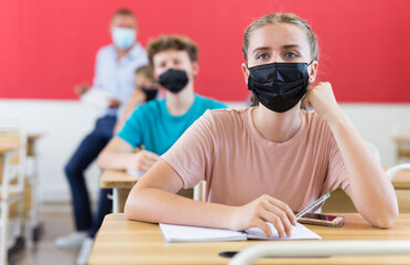 Teenagers in face masks sitting in class room. Male teacher explaining something to them.