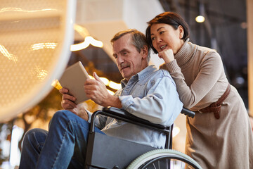 Smiling aged man with mustache sitting in wheelchair and using app on tablet while his Asian wife...