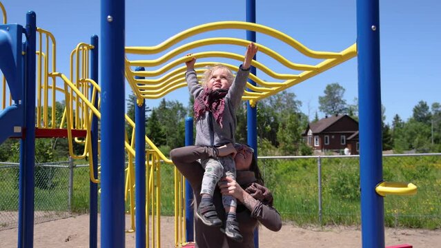 Slow motion video from the front as happy mother lifts two year old son up, so he can reach the tall monkey bars of a climbing frame on a park.