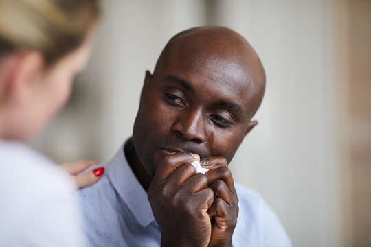Close-up of depressed young bald black man with sad eyes holding napkin in hands and crying while talking to therapist at therapy session