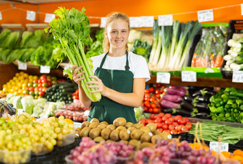 Portrait of a positive fifteen-year-old girl who works part-time in a store as a trainee saleswoman, standing in the ..vegetable department, holding celery in her hands