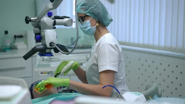 Side view concentrated dental assistant using curing light for polymerization of filling material. Slim confident Caucasian woman sitting in hospital with patient on dental chair at microscope
