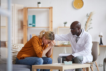 Understanding young Black psychiatrist in white coat and glasses touching shoulder of crying female patient while consoling her at therapy session