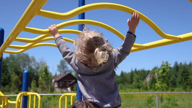Closeup slow mo video as a two year old boy with long blonde hair is seen from behind, stretching up to reach yellow monkey bars on playground.