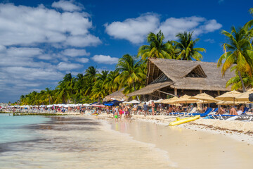 People sunbathing on the white sand beach with umbrellas, bungalow bar and cocos palms, turquoise...