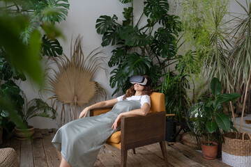 Fototapeta na wymiar Young female wearing virtual reality goggles sleeping in armchair after using VR technology, relaxed woman in AR glasses resting in beautiful indoor tropical garden after watching 3d video