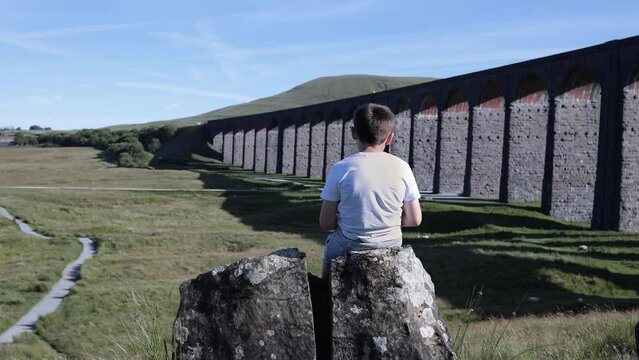 Boy sitting on a stone looking out over a large arched railway bridge. 
Ribblehead Viaduct or Batty Moss Viaduct.