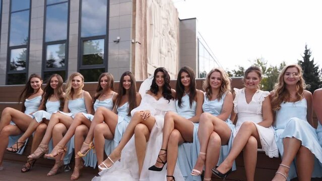 Attractive bride and bridesmaids switching crossing legs sitting on bench seductively in provocative way. Pretty women in identical dresses cross legs simultaneously together flirting at camera.