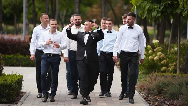 Groom flexing and having fun walking with groomsmen on wedding day. Happy classy man in black suit, bow tie and boutonniere hanging out partying with friends and cheering posing at camera in a park.