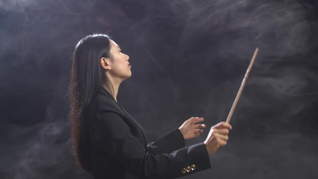 Side View Of Asian Conductor Woman Holding A Baton Closing Her Eyes And Showing Gesture In The Black Studio With Fog 
