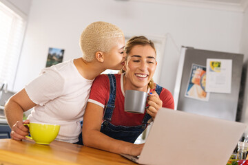 Image of happy diverse lesbian couple watching laptop and drinking coffee