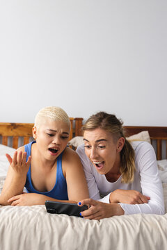 Vertical Image Of Suprised Diverse Lesbian Couple Using Smartphone In Bed