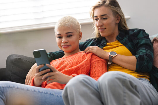 Image of happy diverse lesbian couple sitting on sofa and using smartphone
