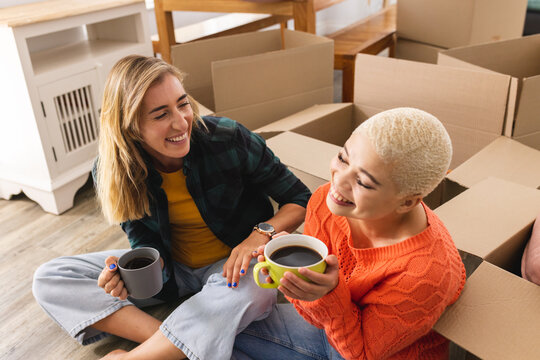Image of happy diverse lesbian couple drinking coffee in new house