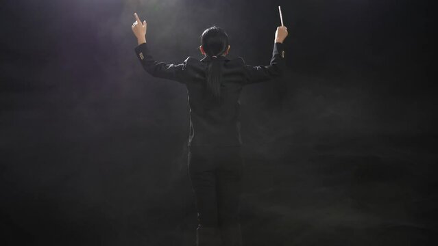Back View Of Asian Conductor Woman Holding A Baton And Showing Gesture In The Black Studio With Fog 
