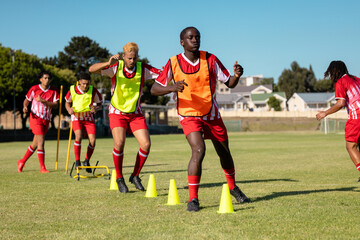 Multiracial male players in red jerseys running around yellow cones at playground against clear sky