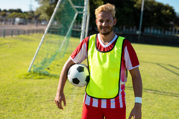 Portrait of smiling caucasian young player in red jersey with soccer ball standing in playground