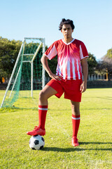 Portrait of biracial young male athlete wearing red jersey with soccer ball standing on grassy land