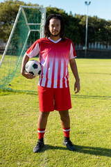 Obraz premium Confident biracial male player with dreadlocks in red jersey holding ball and standing on grass