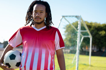 Serious biracial male player with dreadlocks wearing red jersey with ball standing in playground