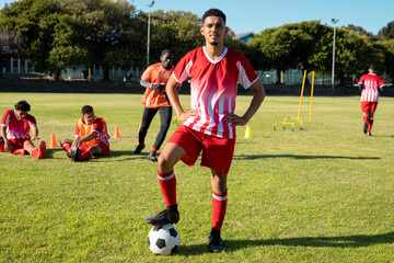 Multiracial confident male player in uniform with arms akimbo and leg on soccer ball at playground