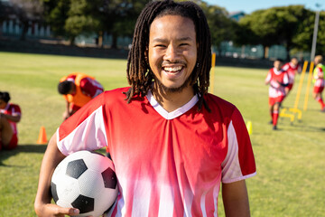 Portrait of biracial male player with dreadlocks holding soccer ball and laughing at playground