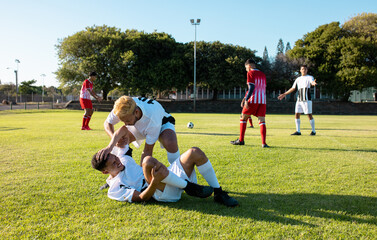 Multiracial male player assisting injured teammate lying on field in getting up during match