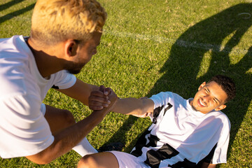 High angle view of caucasian player assisting injured teammate lying on field in getting up