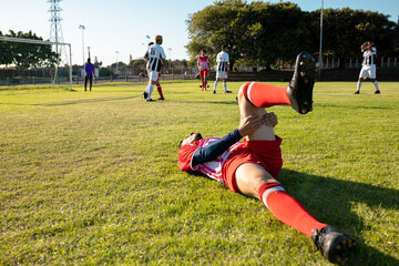 Multiracial injured player lying on grassy field and teammates playing in background during match