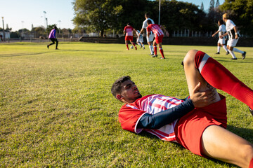 Fototapeta premium Multiracial player with physical injury lying on grassy field and teammates playing in background