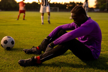 Obraz premium Side view of sad african american male goalkeeper sitting on field at playground during sunset