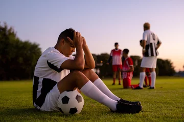 Fotobehang Side view of caucasian sad male player with head in hands and ball sitting on field at sunset © wavebreak3