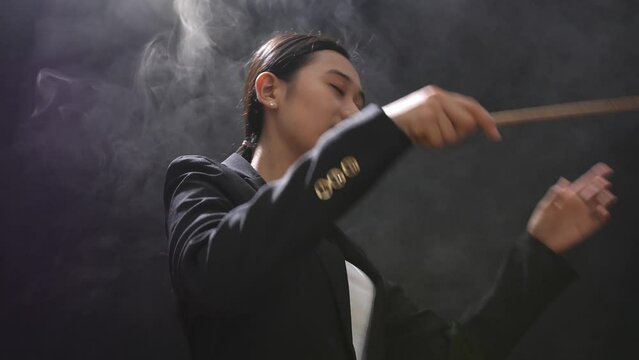Side View Of Asian Conductor Woman Holding A Baton And Showing Gesture Quickly In The Black Studio With Fog 
