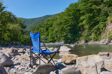 A blue folding chair stands on a boulder near a mountain river. You can see forest, mountains and...