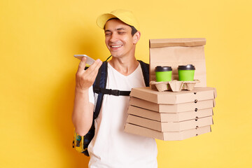 Smiling man courier in white T-shirt and cap isolated over yellow background, holding pizza and...