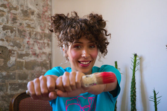 Young Beautiful Latina And Artist Holding A Paintbrush In Her Hands Showing It To The Camera While Doing Different Poses And Smiling For The Photo. Empowered Woman Concept