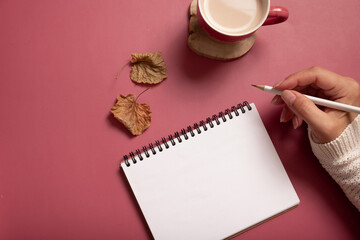 Obraz na płótnie Canvas Table desk with notepad and female hand wit pencil and fall leaves on burgundy colored background. Autumn flat lay composition
