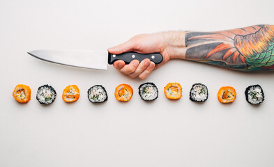 Japanese rolls and a man's hand with a knife and a colorful bright tattoo on a white background. - 516915779