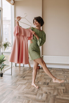 Caucasian brunette woman staying on the room in green costume . Full view of young cute fair skinned girl holding pink dress at home. Concept of home girl mood, lifestyle 