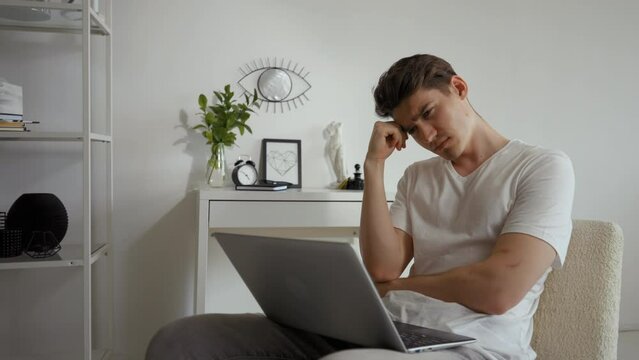 Man feels tired listening to boring online lecture via laptop. Young sleepy student sits on chair in living room yawning and covering mouth with hand