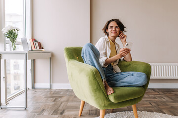 Attractive smiling woman with phone at home. Full view of charming woman holding smartphone sitting on the chair in blue jeans, white shirt. Lifestyle concept