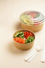 Trendy wholesome dish poke or buddha bowl - rice, wakame seaweed, tomatoes, cucumber, and red fish salmon - in a recycled round carton with sustainable fork, takeaway food concept on beige background