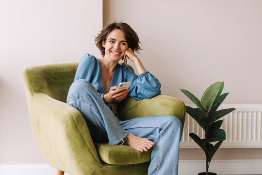 View of beautiful woman in home with phone .Brunette carre hair look and smiling to camera sitting on the green chair at home. Relax, lifestyle concept 