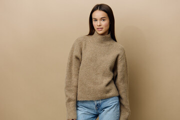 a beautiful, young, attractive woman with long, well-groomed hair stands in a warm textured sweater on a beige background, posing with her hands in a relaxed manner and looking affably into the camera