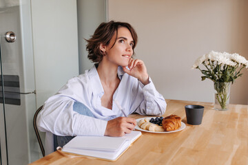 Image of attractive woman looking up and thinking . Brunette short hair woman sitting in jeans and blues shirt at home with notebook, cup coffee, croissant. Concept of working mood 