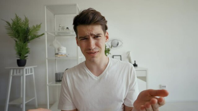 POV of shocked young man grimacing and refusing to look in camera. Guy sits in modern living room at home talking on video call with friend closeup