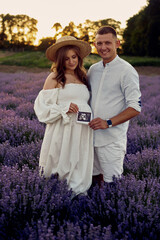 portrait of a young beautiful pregnant couple on a lavender field at sunset holding first ultrasonography image. Happy family concept