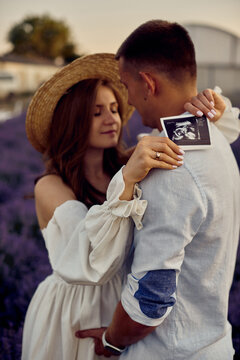 portrait of a young beautiful pregnant couple on a lavender field at sunset holding first ultrasonography image. Happy family concept