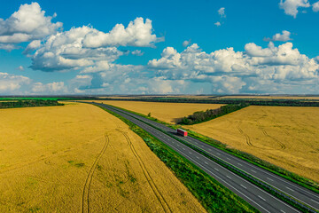 red truck driving on asphalt road along the yellow wheat fields with a cloudy sky. seen from the...